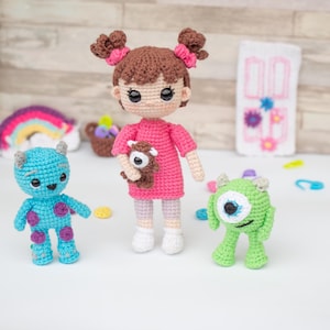 May include: A crocheted doll with brown hair and a pink dress holding a brown teddy bear. Two crocheted monsters, one blue and one green, stand beside the doll.
