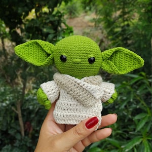 Puede incluir: Un juguete de peluche verde tejido a crochet con orejas grandes y una capa blanca tejida a crochet. El juguete tiene ojos negros y una pequeña nariz.