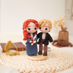 Op de afbeelding: Twee handgemaakte gehaakte poppen die samen op een ronde houten voet staan. De linkerpop heeft lang krullend rood haar, een blauwe jurk en een witte sjaal. De rechterpop heeft kort blond haar, een zwart jasje, een wit overhemd en een bruine broek.