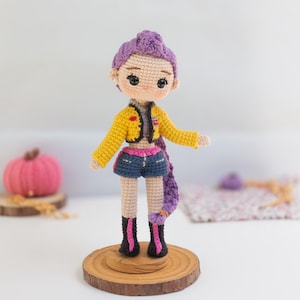 Puede incluir: Muñeca de ganchillo hecha a mano con cabello morado, chaqueta amarilla, pantalones cortos de mezclilla y botas negras con detalles rosas. La muñeca se exhibe sobre una base de madera, mostrando un diseño artesanal único.