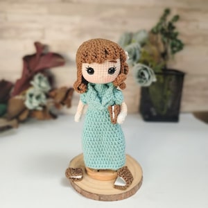 May include: A crocheted doll with brown hair, a light blue dress, and brown shoes. The doll is standing on a wooden base.