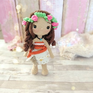 Puede incluir: Una muñeca de crochet con cabello castaño, que lleva una corona de flores, un vestido blanco y naranja y un collar azul. La muñeca está de pie sobre una superficie de madera.
