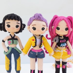 May include: Three handmade crochet dolls with unique outfits and hairstyles. The dolls have black, purple, and pink hair, and wear colorful tops, shorts, and skirts. The dolls are standing side by side, showcasing the craftsmanship of the crochet work.