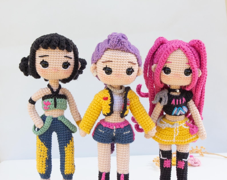 May include: Three handmade crochet dolls with unique outfits and hairstyles. The dolls have black, purple, and pink hair, and wear colorful tops, shorts, and skirts. The dolls are standing side by side, showcasing the craftsmanship of the crochet work.