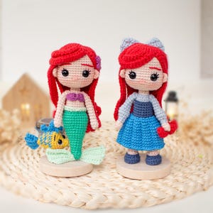 May include: Two crocheted dolls with bright red yarn hair. The left doll has a purple top, a green mermaid tail, and a small yellow and blue fish. The right doll wears a light blue long-sleeved top, a blue skirt, and a light blue hair bow.