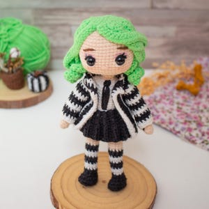 May include: A crocheted doll with green hair, a black and white striped jacket, a black skirt, and black and white striped stockings. The doll is standing on a wooden base.