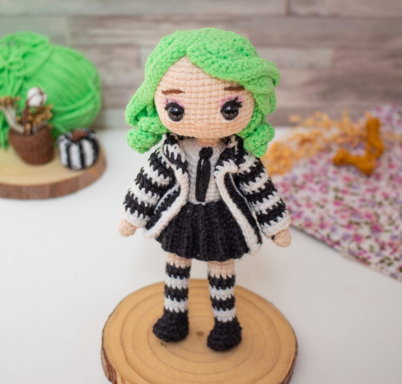 May include: A crocheted doll with green hair, a black and white striped jacket, a black skirt, and black and white striped stockings. The doll is standing on a wooden base.