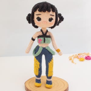 May include: A handmade crochet doll with black hair styled in two buns. The doll wears a green and blue top, blue pants with yellow accents, and yellow shoes. The doll is standing on a wooden disc.