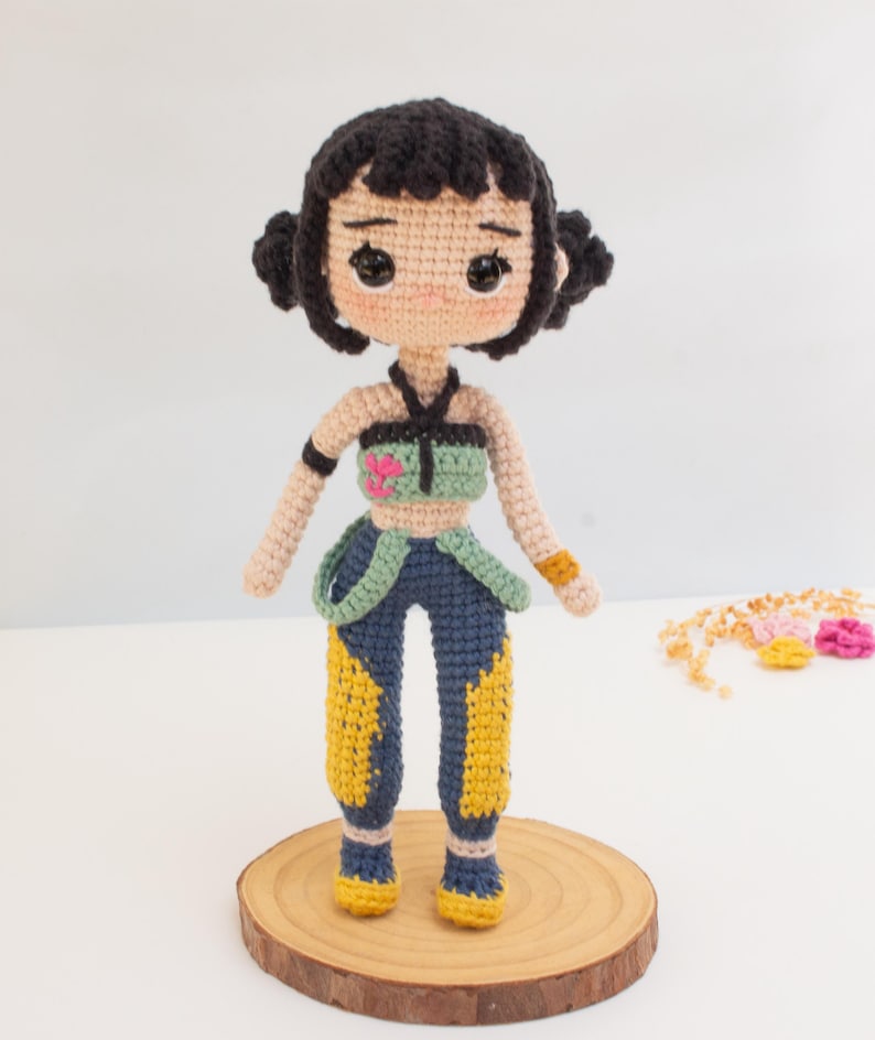 May include: A handmade crochet doll with black hair styled in two buns. The doll wears a green and blue top, blue pants with yellow accents, and yellow shoes. The doll is standing on a wooden disc.
