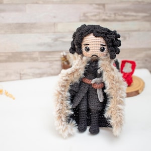 Könnte beinhalten: Eine handgefertigte Häkelpuppe, die eine Figur mit lockigem schwarzem Haar, großen schwarzen Augen und einem kleinen Mund darstellt. Die Figur trägt ein schwarzes Outfit mit braunem Gürtel, ein graues Schwert und einen langen, beigen Kunstfellumhang.