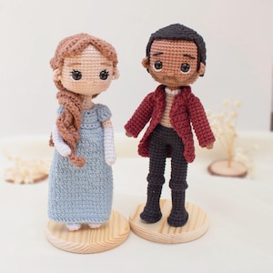 May include: Two crocheted dolls, a woman in a blue dress and a man in a red coat, stand on wooden bases. The woman has brown hair in a braid, and the man has black hair. The dolls are handmade and have detailed features.