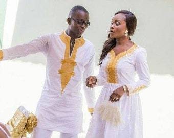 May include: A couple wearing matching white and gold traditional African clothing. The man is wearing a long-sleeved shirt with gold embroidery and the woman is wearing a dress with gold embroidery.