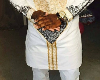 Op de afbeelding: Een wit overhemd met lange mouwen met gouden en zwarte geborduurde patronen. Het overhemd heeft een kraag en een lang, vloeiende ontwerp. Het overhemd wordt gecombineerd met een witte broek en gouden tassel loafers.