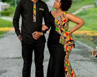 Può includere: Una coppia in abito formale, che cammina su un sentiero lastricato. L'uomo indossa una camicia nera a maniche lunghe e pantaloni con accenti colorati. L'abito nero della donna presenta un design vivace e fantasia sul corpetto e sulla gonna, creando un contrasto sorprendente.