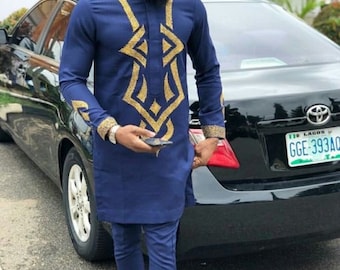 Può includere: Un uomo indossa una camicia blu a maniche lunghe con motivi geometrici dorati, pantaloni blu abbinati e un cappello nero. È in piedi accanto a un'auto nera con una targa che recita "Lagos GGE-393AC".