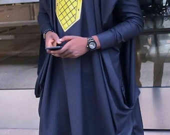 Puede incluir: Un hombre con un atuendo tradicional africano negro y amarillo, con un sombrero negro y gafas de sol. Está de pie frente a una puerta de vidrio y sostiene un teléfono móvil.