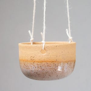 May include: A hanging ceramic planter with a speckled, two-tone design. The upper portion is a light tan color, while the lower half is a speckled, muted purple. White cords are attached for hanging, making it suitable for indoor or outdoor use.