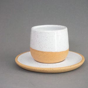 May include: A handmade ceramic cup and saucer set. The cup is white with speckled detailing and a tan base. The saucer is white with speckled detailing and a tan rim. The set is designed for serving beverages.
