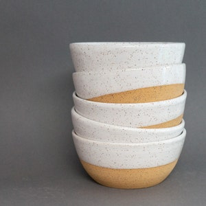 May include: A stack of five ceramic bowls. The top half of each bowl is white with speckled detailing, while the bottom half is a light tan color. The bowls are stacked on top of each other against a gray background.