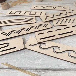 May include: A set of nine wooden stencils featuring various patterns, such as wavy lines, zigzags, and straight lines. The stencils are crafted from light brown wood and are displayed on a white surface.