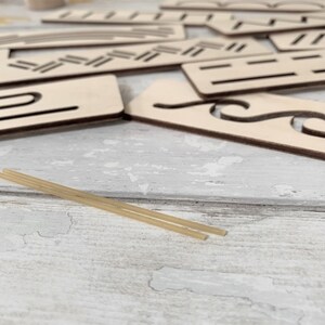 May include: A set of wooden stencils with various shapes and patterns, including wavy lines, zigzags, and a curved shape. Two thin, light brown sticks are lying on a white wooden surface.