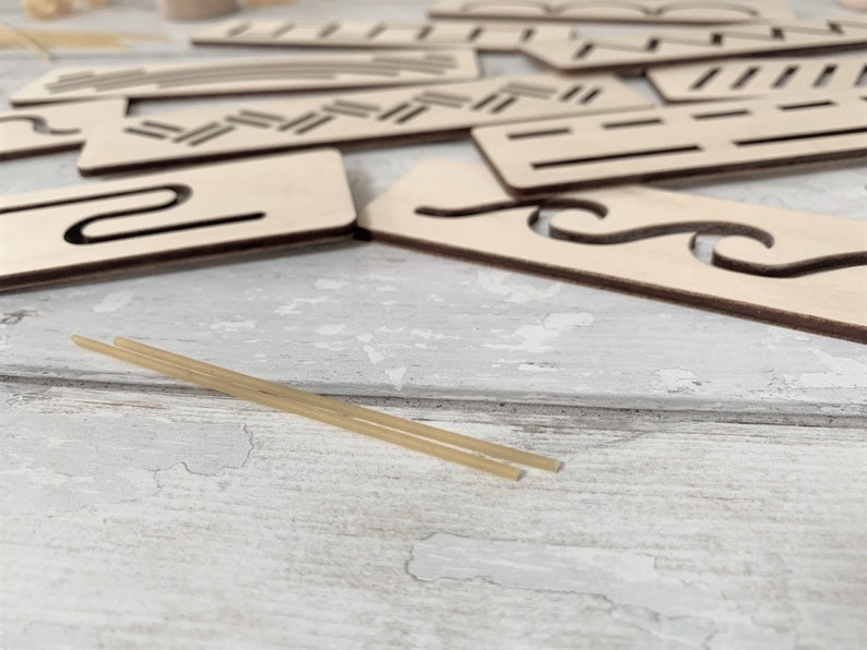 May include: A set of wooden stencils with various shapes and patterns, including wavy lines, zigzags, and a curved shape. Two thin, light brown sticks are lying on a white wooden surface.
