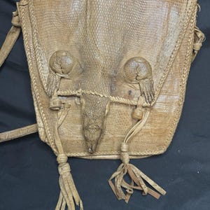 May include: Tan leather crossbody bag with a reptile-like texture. The bag features a flap closure with two decorative knobs and a central, stylized snout. Tassels hang from the bottom, and the bag has a long strap.
