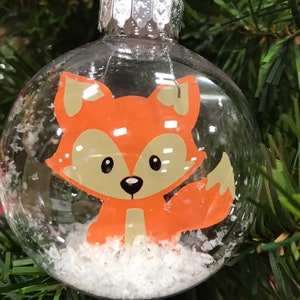 May include: Clear glass Christmas bauble with a white and orange cartoon fox inside. The bauble is filled with white glitter resembling snow.