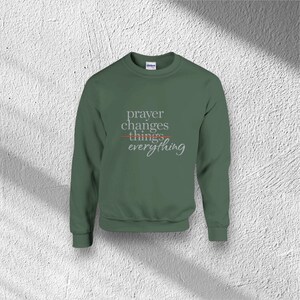 Puede incluir: Una sudadera verde con el texto "prayer changes things everything" en una fuente estilizada. La palabra "things" est&aacute; tachada con una l&iacute;nea roja. La sudadera es de color liso y tiene cuello redondo.