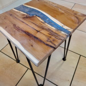 May include: A small, square wooden side table with a live edge and a blue resin river. The tabletop is supported by four black hairpin legs. The wood has a natural, warm brown tone.