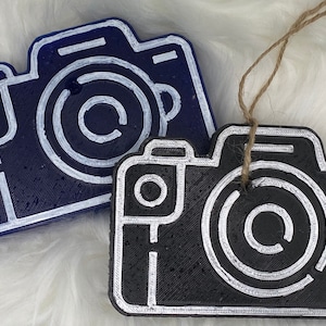 May include: Two camera-shaped ornaments, one black with white details and the other blue with white details. Both have a string attached for hanging.