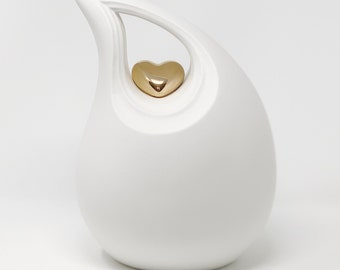 May include: A white ceramic urn with a teardrop shape and a gold heart-shaped accent. The urn has a smooth, glossy finish and a curved handle-like opening at the top. The gold heart is centered within the opening.