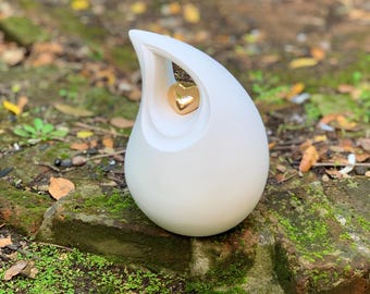 May include: A white teardrop-shaped urn with a gold heart-shaped accent. The urn has a smooth, glossy finish and a unique design. The background features a natural outdoor setting with greenery and fallen leaves.