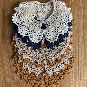 Könnte beinhalten: Eine Auswahl handgefertigter Häkelkragen in Creme, Marineblau und Senfgelb. Jeder Kragen hat ein zartes, spitzenartiges Muster und wird mit kleinen, perlmuttartigen Knöpfen geschlossen. Die Kragen sind auf einer Holzoberfläche angeordnet.