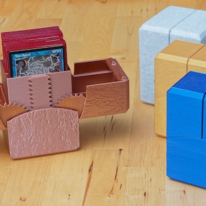 Könnte beinhalten: Vier verschiedene farbige Kartenspiel-Deckboxen. Die Boxen sind aus Kunststoff und haben eine strukturierte Oberfläche. Die Boxen sind geöffnet und zeigen das Innere der Boxen. Eine Box ist roségold, eine ist gold, eine ist hellblau und eine ist dunkelblau.