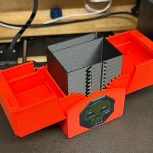 May include: A 3D-printed storage box with a hinged lid and two side compartments. The box is primarily bright red with a central grey compartment. A green and black skull emblem is on the front.
