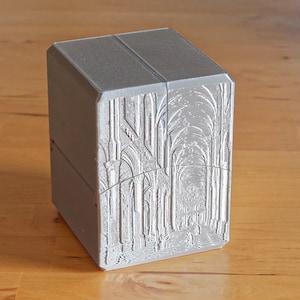 Peut inclure: Une boîte rectangulaire grise avec un motif en relief détaillé d'un couloir voûté. Le motif est en nuances de gris et de blanc.