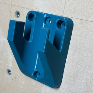 Könnte beinhalten: Eine türkisblaue, 3D-gedruckte Halterung mit einer einzigartigen Form, die zwei vertikale Schlitze und mehrere Befestigungslöcher aufweist. Die Halterung ist auf einer hellen Oberfläche mit sichtbaren Schrauben montiert.