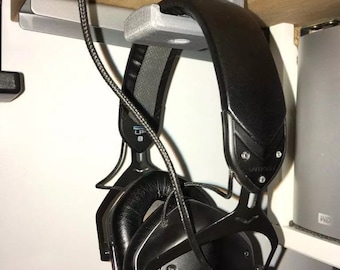 May include: A grey 3D printed headphone hook mounted under a wooden desk. A pair of black headphones are hanging on the hook.