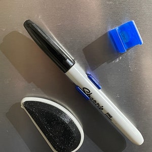 May include: A black dry-erase marker with a white barrel and blue accents. The marker says "Sharpie Fine Point" on the barrel. There is a blue plastic scraper and a black and white eraser on a silver surface.