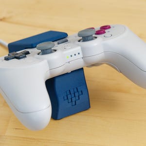 May include: A white wireless video game controller with blue and purple buttons, resting on a blue charging stand. The stand has a pixelated heart design.