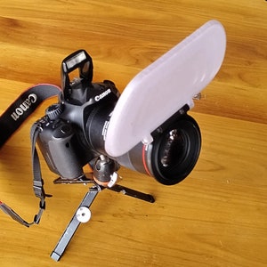 Peut inclure: Un appareil photo reflex numérique Canon noir avec un objectif et un diffuseur blanc sur l'objectif. L'appareil photo est monté sur un petit trépied noir.