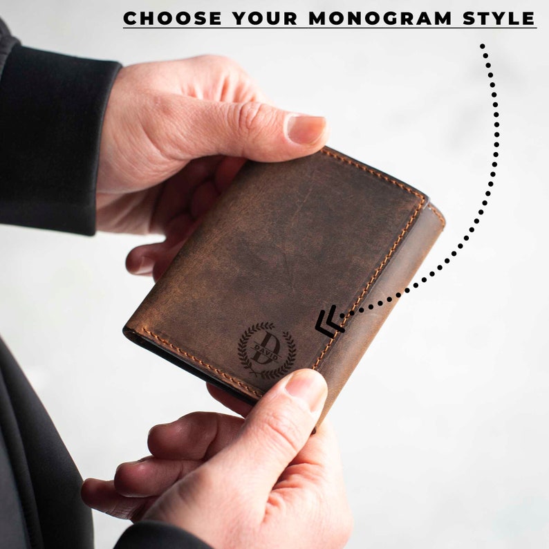 May include: A brown leather wallet with a monogrammed initial "D" in a laurel wreath design. The wallet is folded in half and held in a hand.