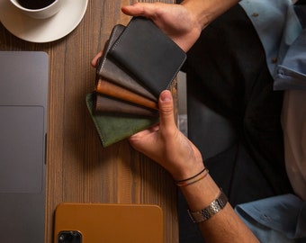 May include: A person holding a stack of five leather wallets in different colors, including black, brown, green, and gray. The wallets are arranged in a fan shape, with the black wallet on top. The person is wearing a blue shirt and a silver bracelet.