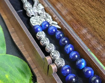 Peut inclure: Un chapelet composé de perles en œil de tigre bleu poli et d'entretoises en métal argenté ornementées. Le chapelet est présenté dans une boîte rectangulaire en bois avec une doublure intérieure sombre, incluant un pompon métallique.