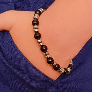 May include: A black beaded bracelet with silver-colored spacers. The bracelet is made of round, polished black beads and small, cylindrical silver-toned metal beads. The bracelet is worn on a wrist, with a blue background.