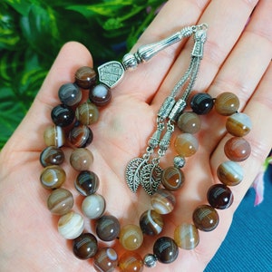 May include: A prayer bead bracelet with brown and white striped agate beads. The bracelet has a silver-toned metal divider and tassel with leaf-shaped charms. The beads are round and vary in shades of brown, tan, and white.