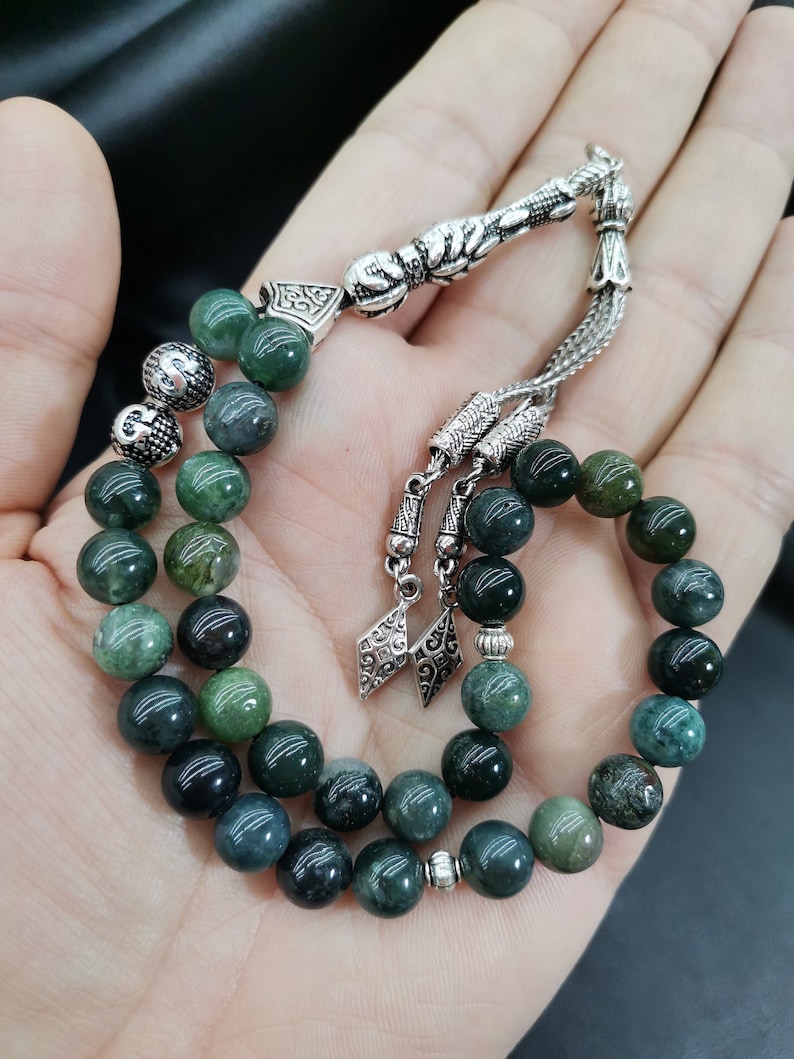 May include: A prayer bead set with dark green and silver accents. The beads are round and have a marbled appearance. The silver components include decorative caps, a connecting piece, and a tassel. The beads are held in an open hand.