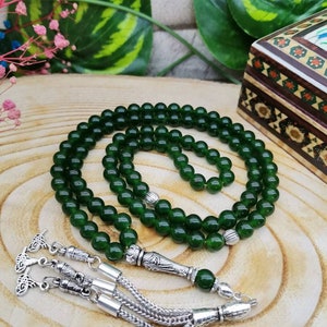 May include: A dark green beaded prayer rosary with silver-colored metal accents. The beads are round and evenly spaced. The rosary is displayed on a wooden surface, with a decorative box in the background.