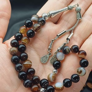 May include: A prayer bead set featuring alternating black, brown, and white agate beads. The beads are strung on a silver chain with decorative silver accents. The beads are held in a hand.